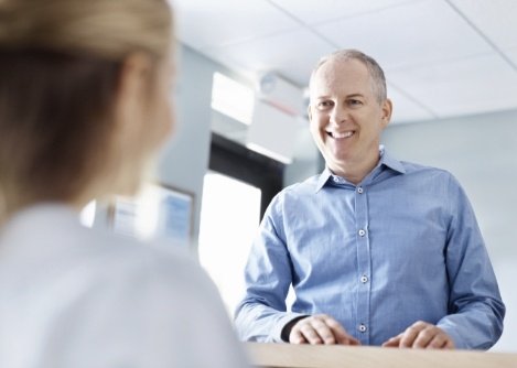 Older man in light blue dress shirt talking to dental team member at front desk